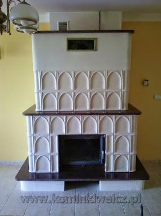 Biały, dwupoziomowy kominek kaflowy z ciemnym blatem, zdobiony łukowymi wzorami, na tle żółtej ściany. Widoczny fragment lampy sufitowej.