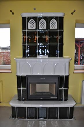 Czarno-biały, kaflowy kominek z żeliwnym wkładem, z dekoracyjnymi elementami i widocznym adresem strony internetowej na tle żółtej ściany.