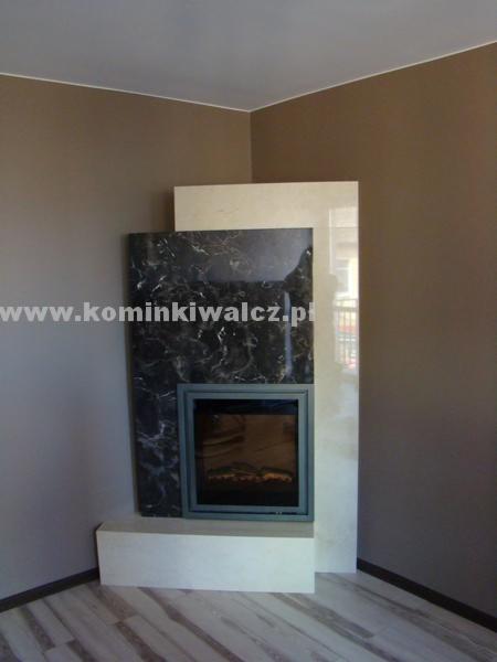 Nowoczesny kominek z czarnym marmurem i jasnym obramowaniem, osadzony w brązowym wnętrzu. Widoczna tekstura marmuru i płomień w palenisku.
