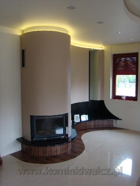 Nowoczesny kominek cylindryczny z czarną obudową i podświetleniem LED w jasnym salonie z drewnianymi akcentami podłogowymi i oknem z żaluzjami.