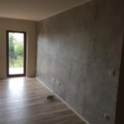 Pok&oacute;j po remoncie: teksturalna szara ściana z gniazdkami, drewnopodobna podłoga, okna z widokiem na zieleń. Nowoczesne wykończenie wnętrza, jasne światło.