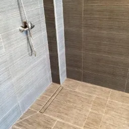 Wykończona kabina prysznicowa z szarymi i brązowymi płytkami imitującymi drewno, z odpływem liniowym w podłodze i chromowaną armaturą.