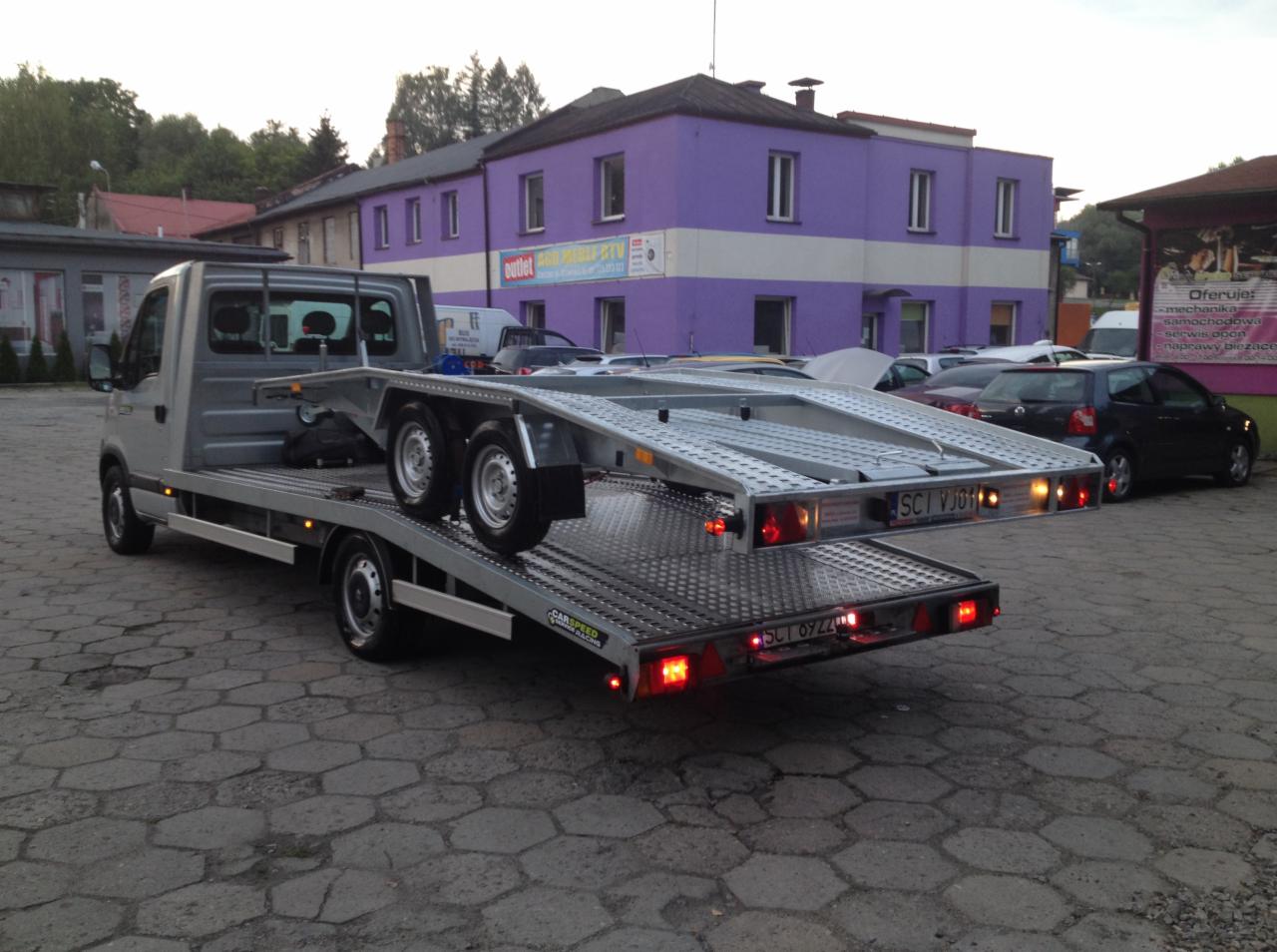 Szara laweta z przyczepą na tle fioletowego budynku. Przyczepa z tablicą rejestracyjną, światła stopu włączone. Widok z tyłu, kostka brukowa na ziemi.