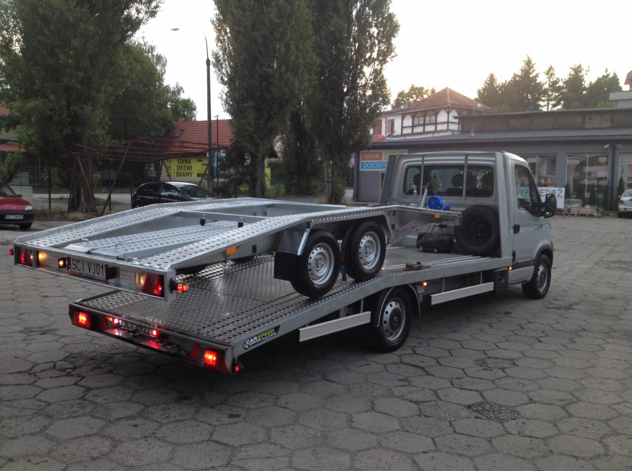 Srebrna laweta z platformą CARSPEED na parkingu, widok z tyłu. Widoczne światła, koła i konstrukcja platformy. Tło: budynki i drzewa.