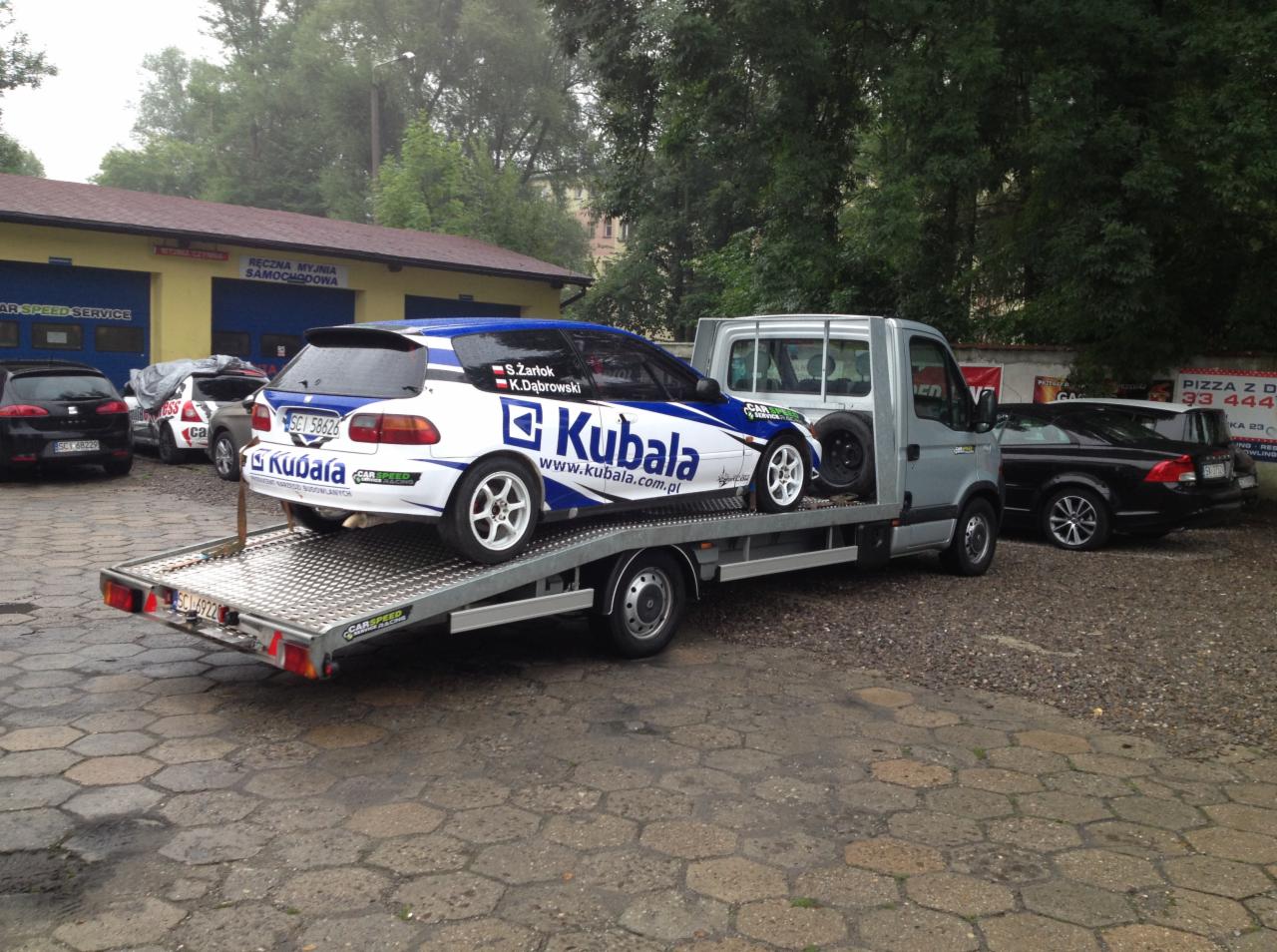Laweta przewozi biało-niebieski samochód wyścigowy Kubala na platformie. W tle budynek myjni samochodowej i inne zaparkowane auta.