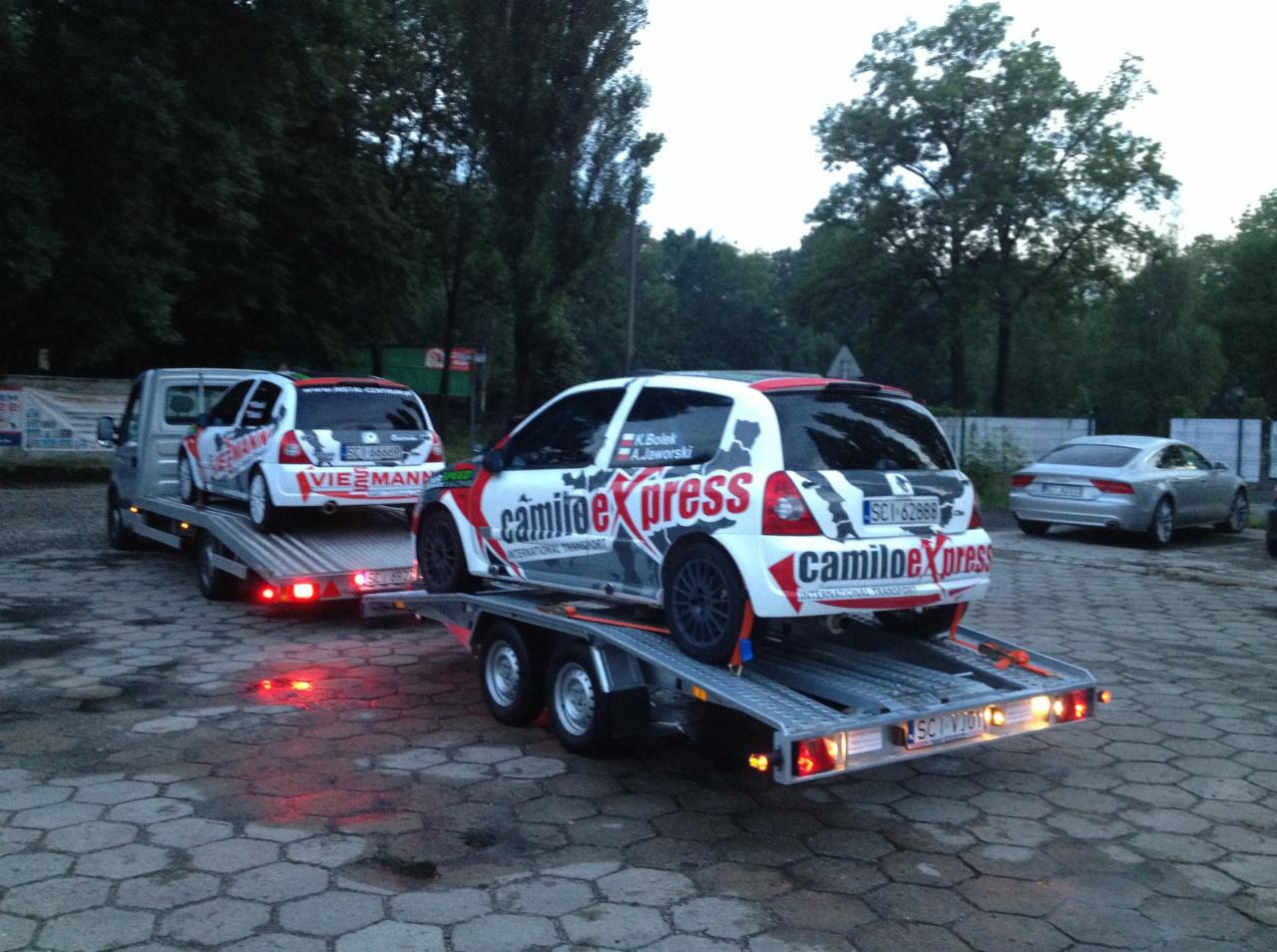 Laweta przewozi dwa samochody wyścigowe z logo Camilo Express. Widoczny srebrny samochód osobowy w tle. Całość na tle zieleni.