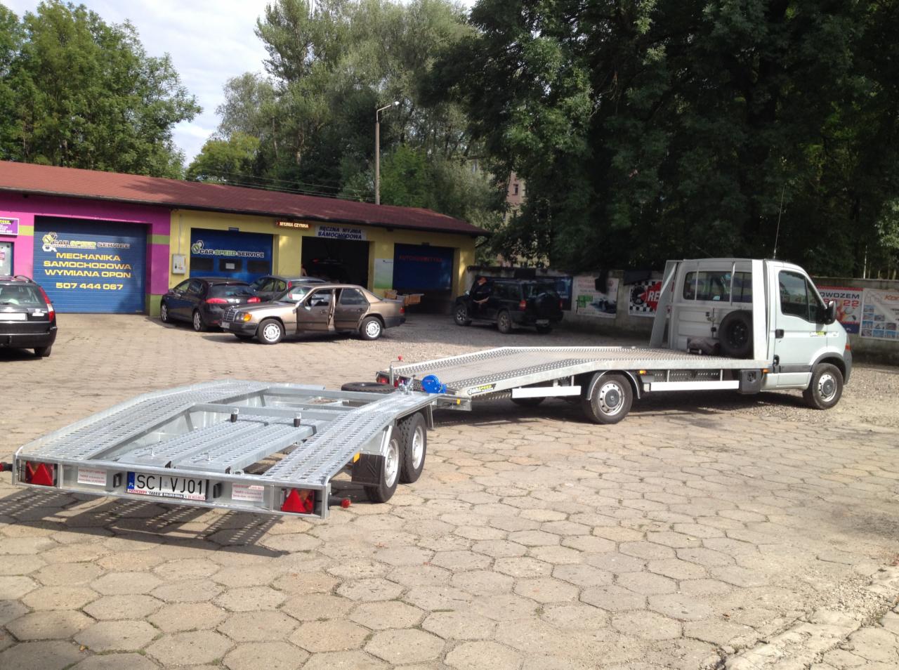 Laweta z przyczepą na betonowym placu, w tle budynek warsztatu samochodowego. Przyczepa posiada tablicę rejestracyjną 'SCI VJ01'.