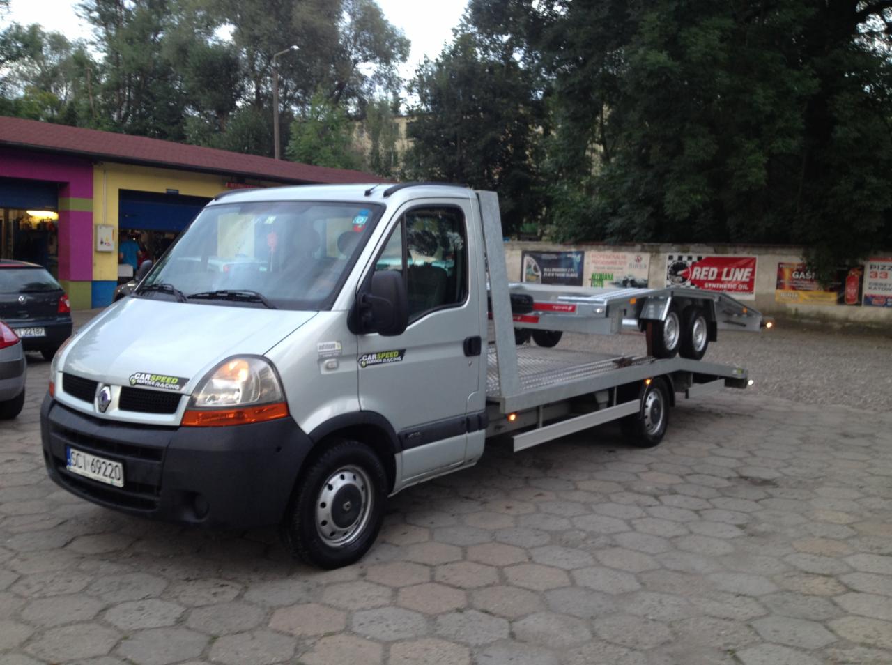 Srebrny samochód-laweta Renault Master z platformą i przyczepką, zaparkowany na brukowanym placu, z budynkiem i drzewami w tle. Widoczne reklamy na murze.
