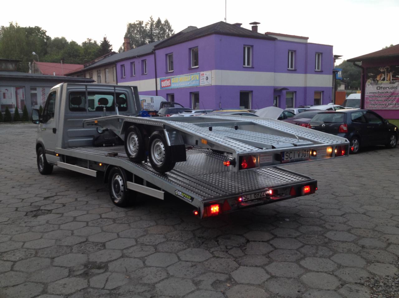 Szara laweta z platformą załadunkową na tle parkingu i fioletowego budynku z napisem 'outlet AGRF'. Widoczne tablice rejestracyjne. Niewyraźne otoczenie.