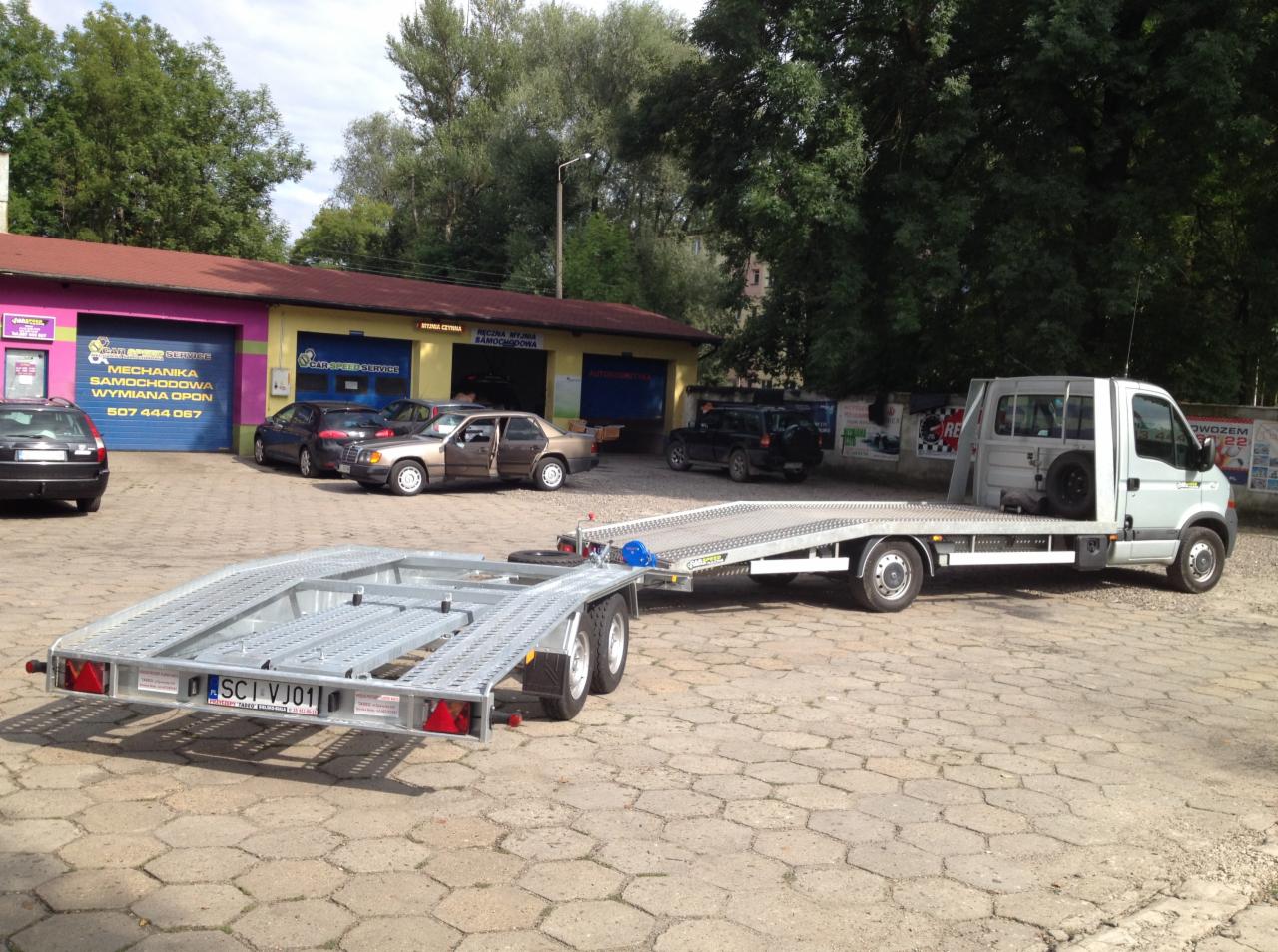 Laweta z przyczepą na tle warsztatu samochodowego w Cieszynie. Widoczne zaparkowane auta i budynek z napisem 'Mechanika Samochodowa Wymiana Opon'.