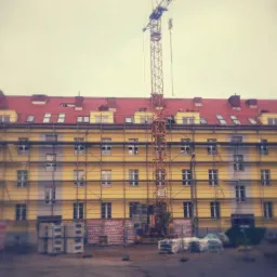 Wielopiętrowy budynek w trakcie remontu, z rusztowaniami i dźwigiem na tle pochmurnego nieba, widoczne materiały budowlane i kontenery na gruz.