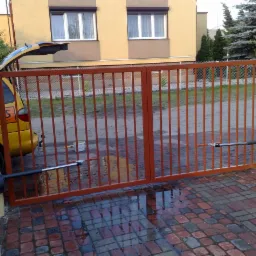 Zewnętrzna dwuskrzydłowa brama w kolorze ceglastym z automatyką, zamontowana na słupkach z żółtego tynku; na mokrym brukowanym podjeździe widoczne odbicia nieba, w tle zaparkowany samochód...