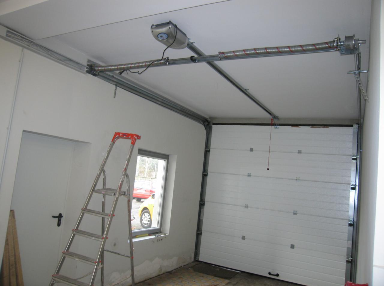 Wnętrze garażu z zamontowaną białą segmentową bramą garażową, widoczny napęd bramy pod sufitem, drabina oparta o ścianę obok okna.