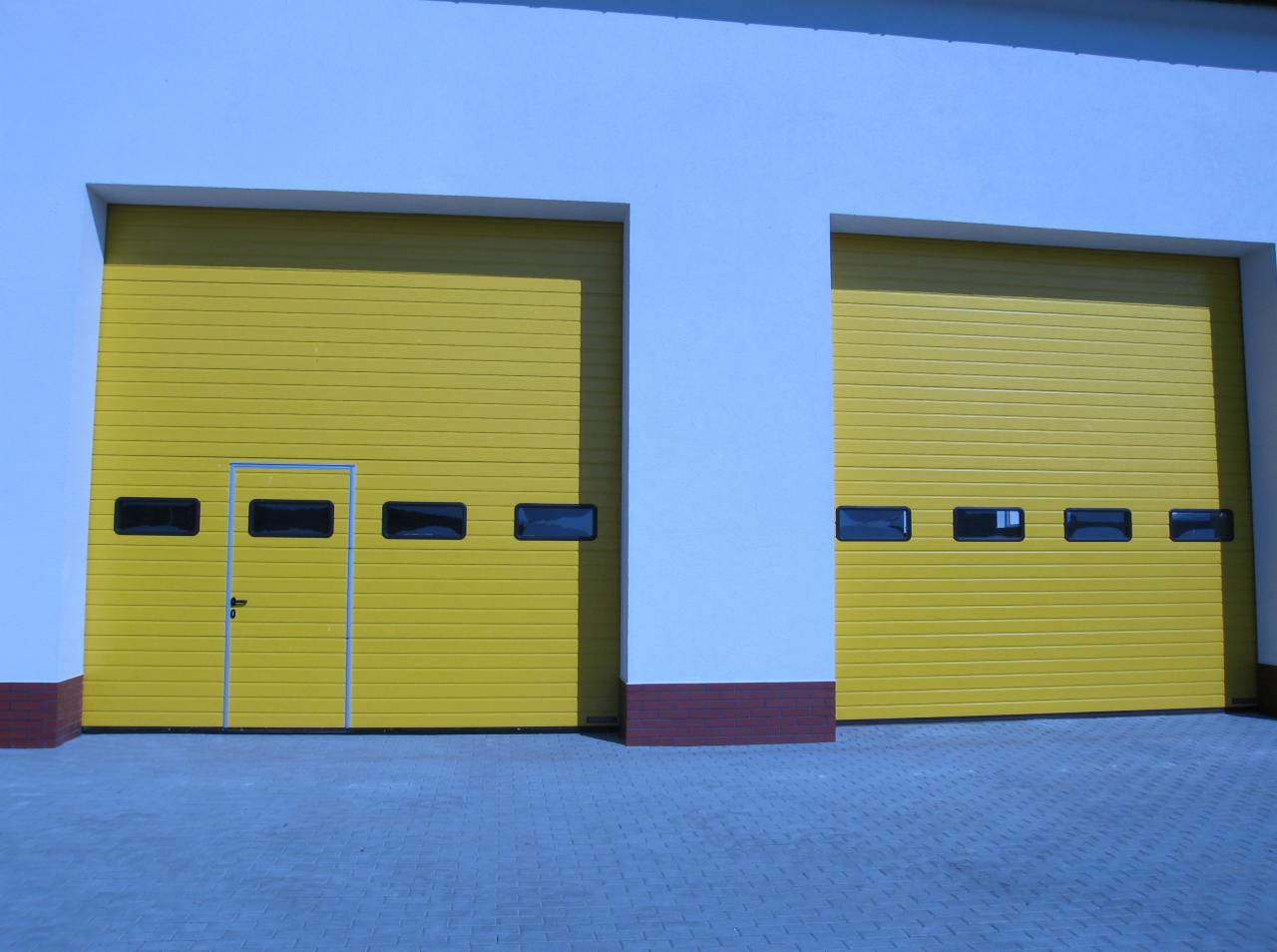 Dwie żółte bramy przemysłowe segmentowe z poziomymi panelami i małymi oknami, jedna z dodatkowymi drzwiami wejściowymi, zamontowane w białej hali z ceglaną podmurówką.