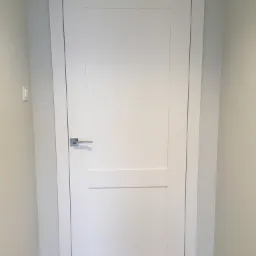 Białe, dwupanelowe drzwi wewnętrzne z prostokątną klamką na tle jasnoszarej ściany i podłogi z parkietu ułożonego we wzór jodełki.