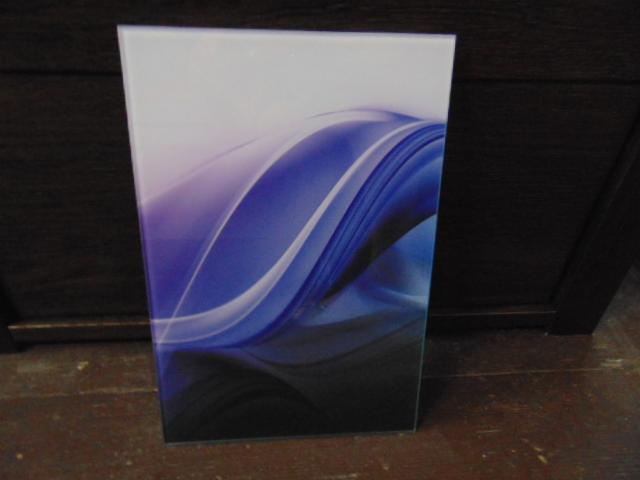 Szklana tafla z abstrakcyjnym motywem fal w odcieniach błękitu, fioletu i bieli, stojąca na drewnianej powierzchni. Gładka powierzchnia szkła odbija światło.