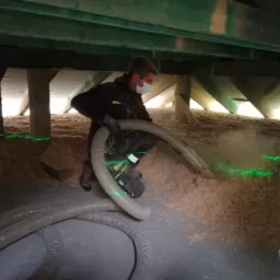 Pracownik w masce i kombinezonie, w trakcie aplikacji izolacji celulozowej na poddaszu, widoczne drewniane elementy konstrukcji dachu i węże do wdmuchiwania izolacji, zielone linie lasera...