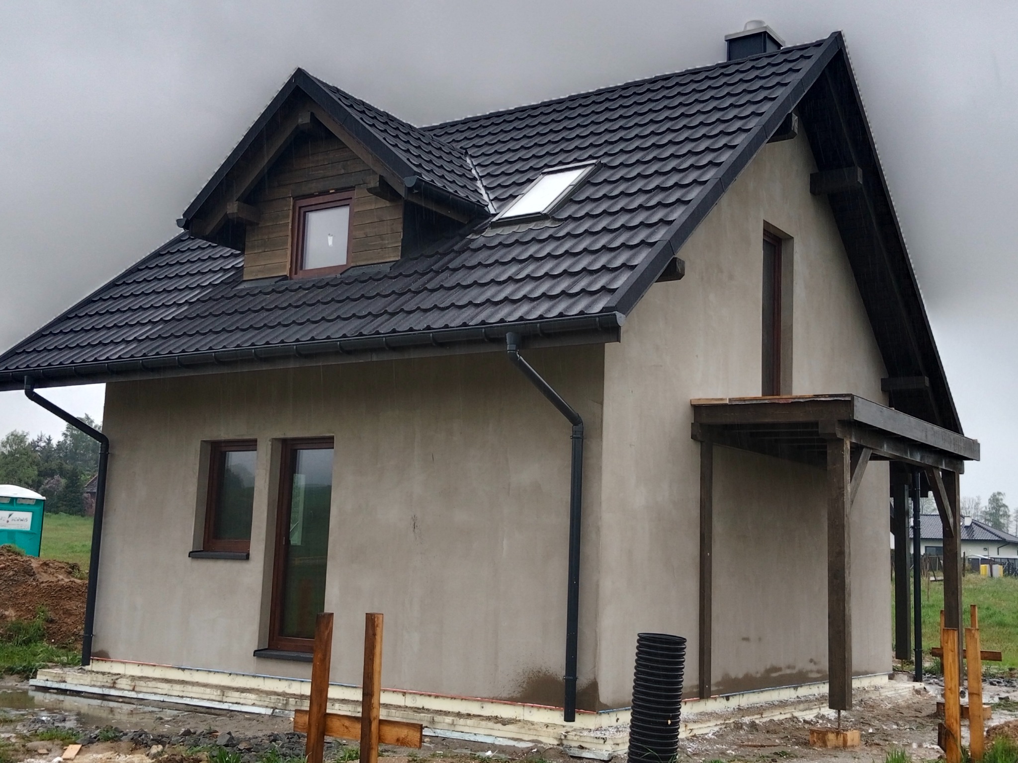Surowy dom szkieletowy z ciemną dachówką, widoczne drewniane elementy konstrukcyjne i szara elewacja w trakcie prac wykończeniowych, otoczenie placu budowy.