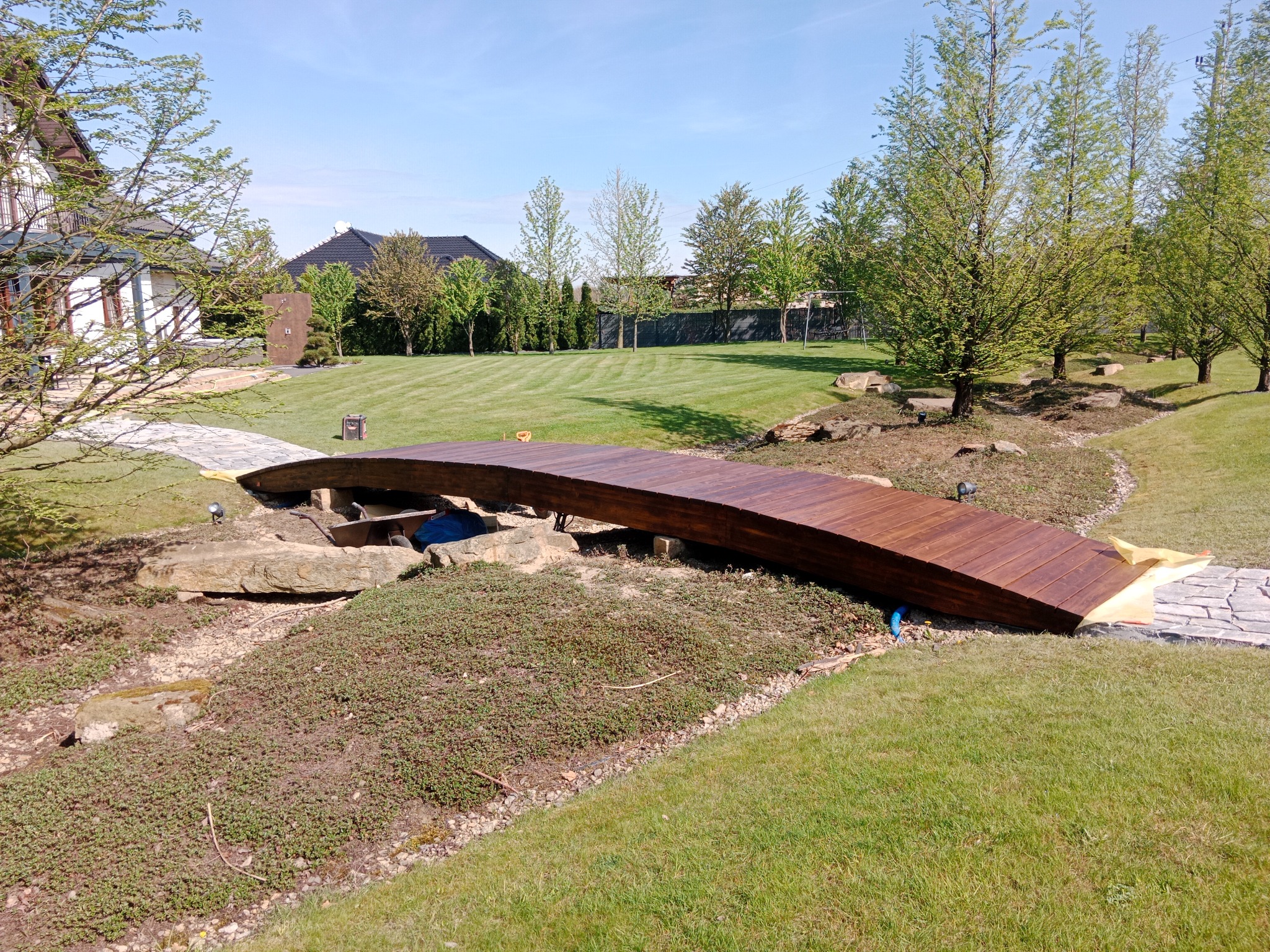 Drewniany, łukowaty mostek nad strumykiem w ogrodzie, z kamiennymi brzegami i ścieżką z płyt.