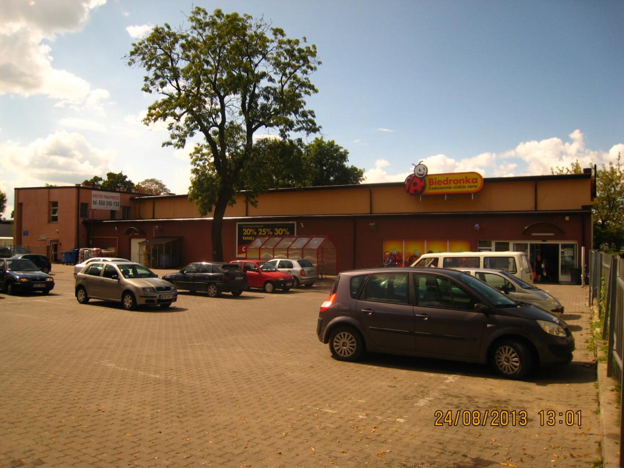 Parking przed budynkiem sklepu Biedronka w słoneczny dzień, widoczne zaparkowane samochody i drzewo.