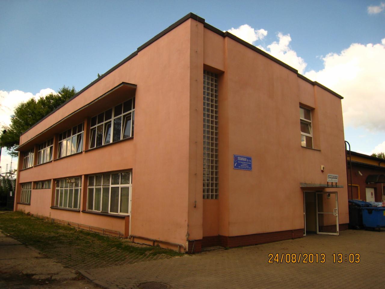 Dwupiętrowy budynek magazynowy w kolorze łososiowym z licznymi oknami, wejściem i tabliczką informacyjną, sfotografowany z zewnątrz w słoneczny dzień.