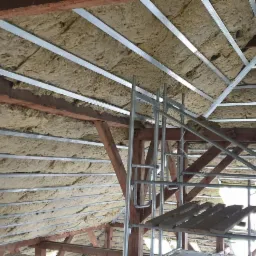 Poddasze ocieplone wełną mineralną, widoczne metalowe profile konstrukcyjne i drewniane elementy więźby dachowej, rusztowanie w tle.