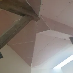 Skośny sufit poddasza z zamontowanymi płytami gipsowo-kartonowymi, widoczne drewniane belki konstrukcyjne i szpachlowane łączenia płyt, w tle okno dachowe i materiały budowlane.