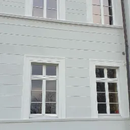 Odnowiona elewacja budynku z czterema białymi oknami z szprosami, widoczne poziome pasy na ścianie, fragment niezagospodarowanego terenu przy podstawie ściany z wystającą rurą.