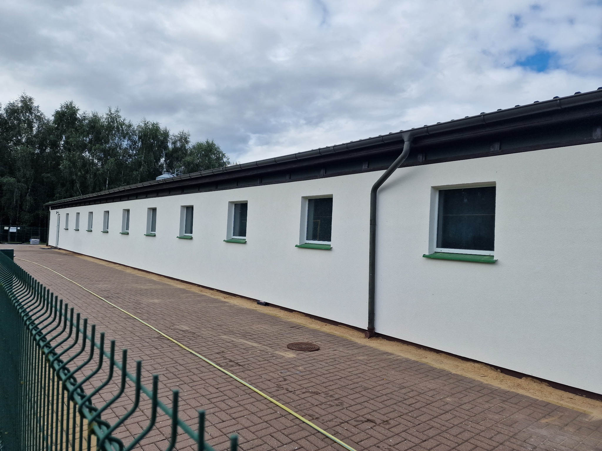 Długi, jednopiętrowy budynek z białymi ścianami i rzędem okien z zielonymi parapetami, widoczny zza zielonego ogrodzenia z siatki, z brązową kostką brukową i żółtym wężem ogrodowym na pierwszym...