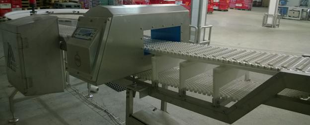 Przemysłowy system detekcji metali z taśmociągiem, widoczny panel sterowania i elementy konstrukcyjne ze stali nierdzewnej, w tle magazyn z paletami.