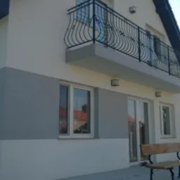 Dwukondygnacyjny dom z balkonem i szarą strefą na elewacji, widok z boku. Ciemna dachówka, białe okna i balustrada z metalu. Na pierwszym planie betonowy taras.