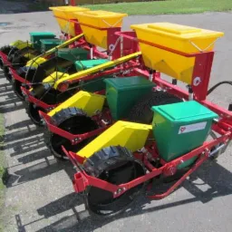 Wielorzędowa siewka punktowa z żółtymi i zielonymi zbiornikami na nasiona, czerwona rama, koła z czarnymi oponami, na betonowej nawierzchni.