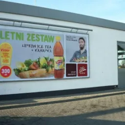 Billboard reklamowy na ścianie budynku prezentujący letni zestaw z Lipton Ice Tea i kanapką, obok wejście do lokalu z szarymi drzwiami.