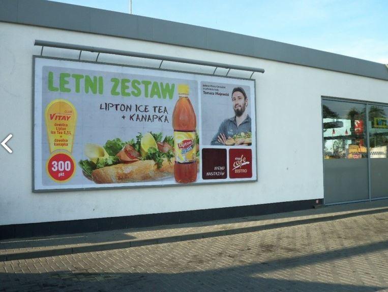 Billboard reklamowy na ścianie budynku prezentujący letni zestaw z Lipton Ice Tea i kanapką, obok wejście do lokalu z szarymi drzwiami.