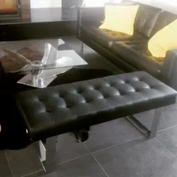 Tapicerowana czarna pufa z pikowaniem i metalowymi nogami w nowoczesnym salonie z czarną skórzaną sofą i żółtymi poduszkami.