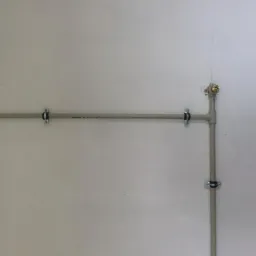 Szare rury instalacyjne zamocowane do białej ściany za pomocą metalowych uchwytów. Widoczne trójniki i zawór. Instalacja na jasnym tle.