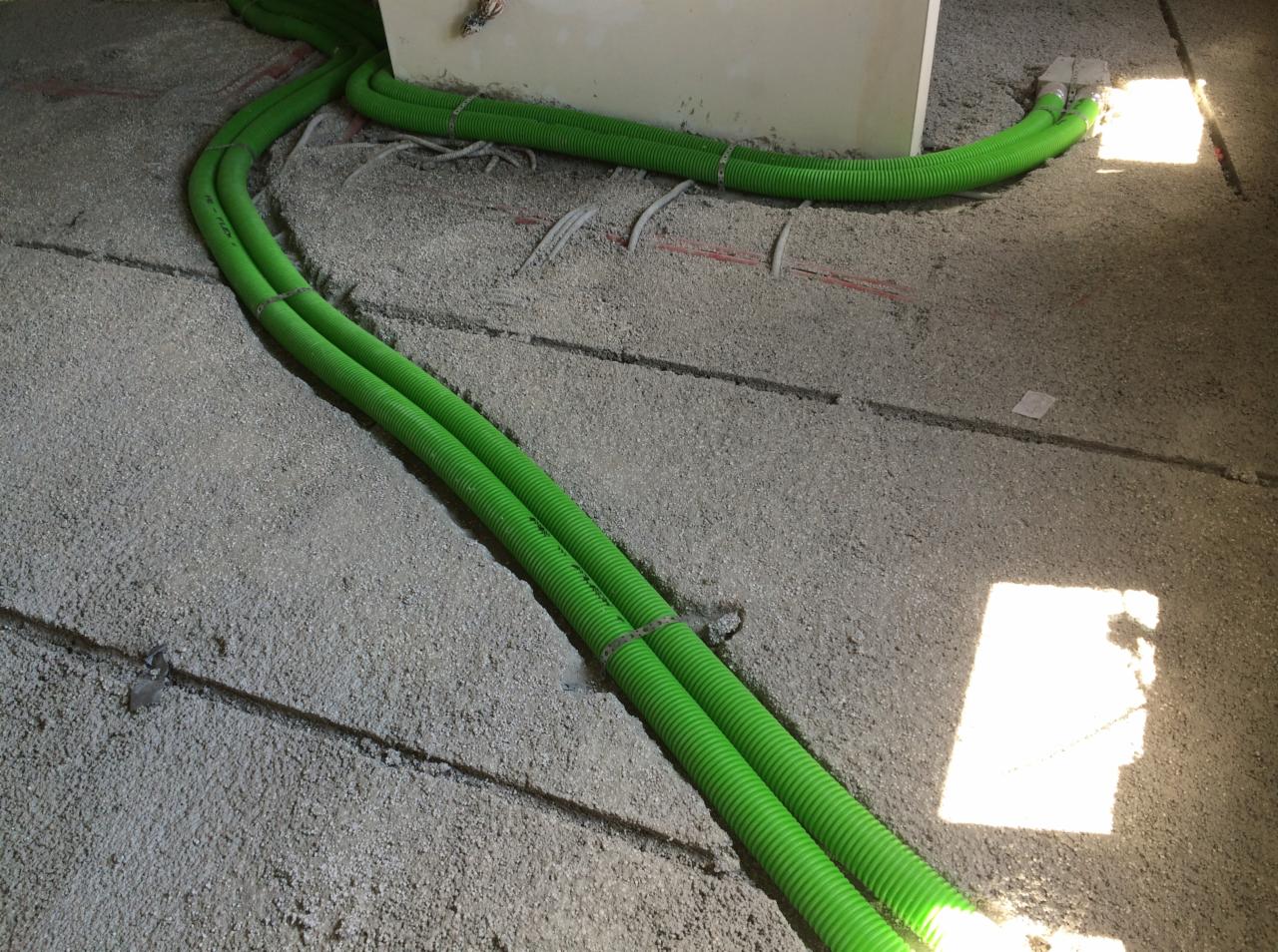 Zielone, karbowane rury wentylacyjne na szarym podłożu z widocznymi bruzdami i elementami instalacji. Fragment ściany w tle. Oświetlenie naturalne.