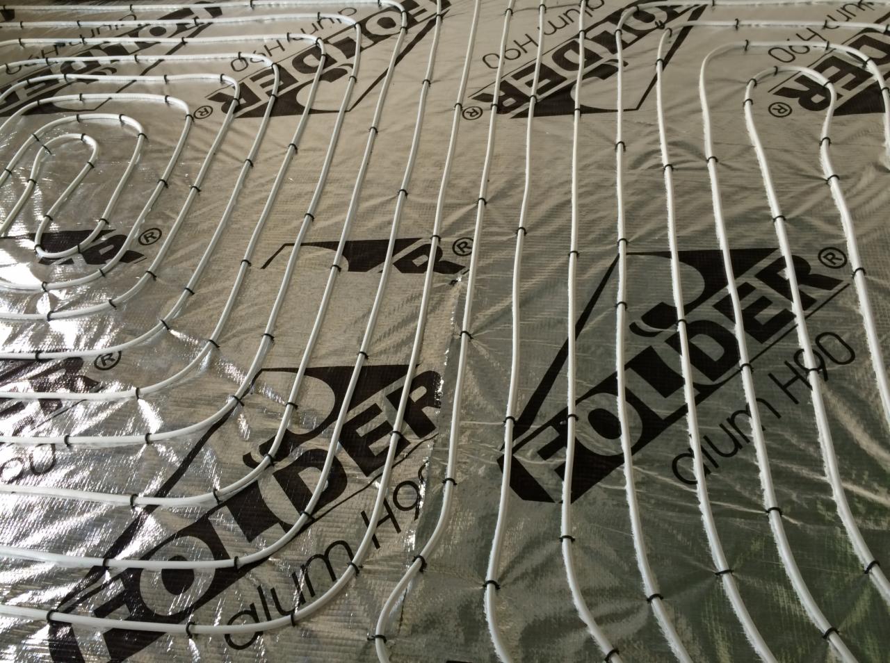 Białe rurki ogrzewania podłogowego ułożone na srebrnej folii z czarnym nadrukiem, przymocowane czarnymi spinkami. Widok z góry, równoległe ułożenie.