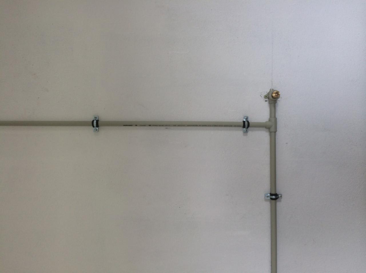 Szare rury instalacyjne zamocowane do białej ściany za pomocą metalowych uchwytów. Widoczne trójniki i zawór. Instalacja na jasnym tle.