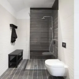 Nowoczesna łazienka z kabiną prysznicową typu walk-in, wykończona białymi i grafitowymi płytkami imitującymi drewno, z wiszącą toaletą i czarnymi akcentami.