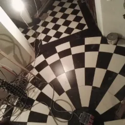 Ułożona podłoga z czarno-białej szachownicy przechodząca w okrągły wzór. Widoczne kable, puszka elektryczna i schody w tle.