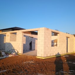 Usługi Ogólnobudowlane Dominik Matyjak - Stan surowy otwarty domu z białego betonu komórkowego w Połajewie, widok z zewnątrz, w tle błękitne niebo. Widoczny pracownik wewnątrz budynku i materiały budowlane.