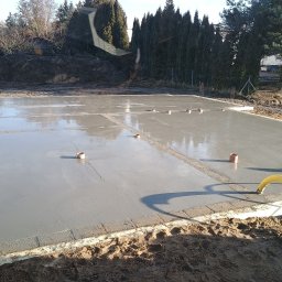 Usługi Ogólnobudowlane Dominik Matyjak - Świeżo wylana, gładka płyta fundamentowa z wystającymi rurami wentylacyjnymi, otoczona szalunkiem. W tle ziemia i drzewa.