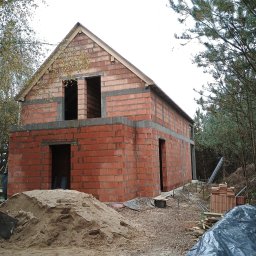 Usługi Ogólnobudowlane Dominik Matyjak - Budowa domu z czerwonej cegły, widoczne otwory okienne i drzwiowe, częściowo zasłonięte stertą piasku. Pracownik z łopatą w tle. Etap murowania ścian.