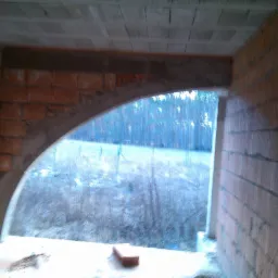 Wnętrze budynku w trakcie budowy: murowane ściany z pustaków ceramicznych, łukowe nadproże okienne z betonu, widok na zewnątrz na łąkę i las. Surowy stan.