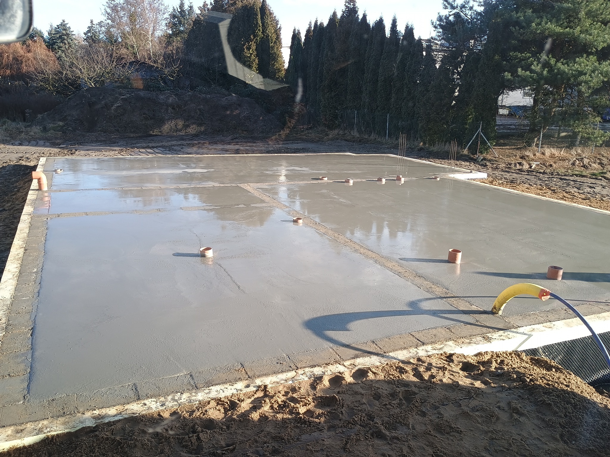 Świeżo wylana, gładka płyta fundamentowa z wystającymi rurami wentylacyjnymi, otoczona szalunkiem. W tle ziemia i drzewa.