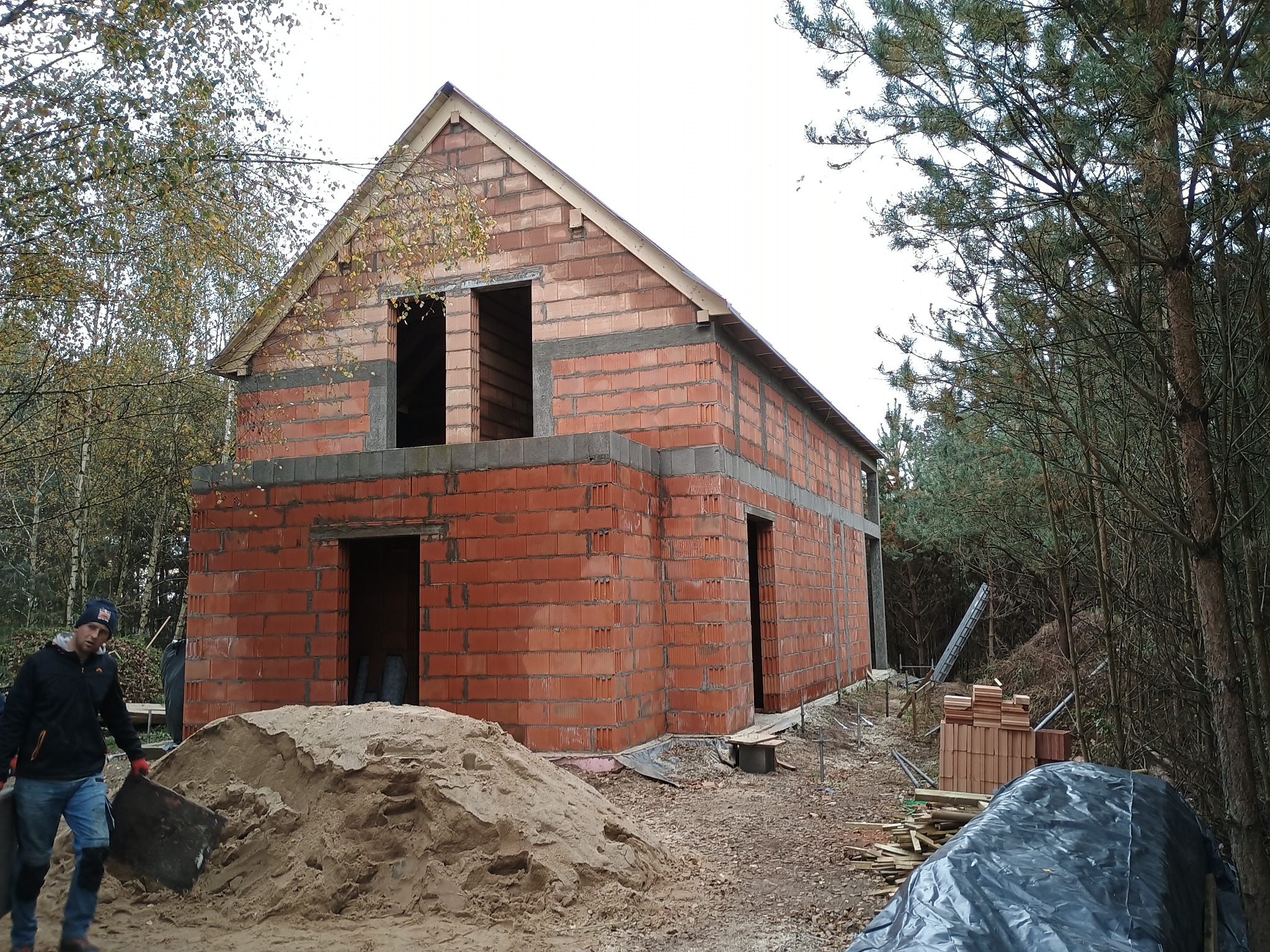 Budowa domu z czerwonej cegły, widoczne otwory okienne i drzwiowe, częściowo zasłonięte stertą piasku. Pracownik z łopatą w tle. Etap murowania ścian.