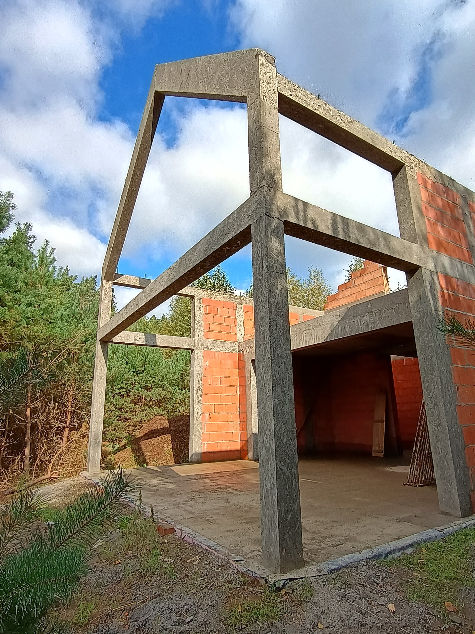 Surowy szkielet domu z betonu komórkowego i cegły, widok z zewnątrz na tle lasu i błękitnego nieba z chmurami. Widać zarys przyszłych okien i dachu.