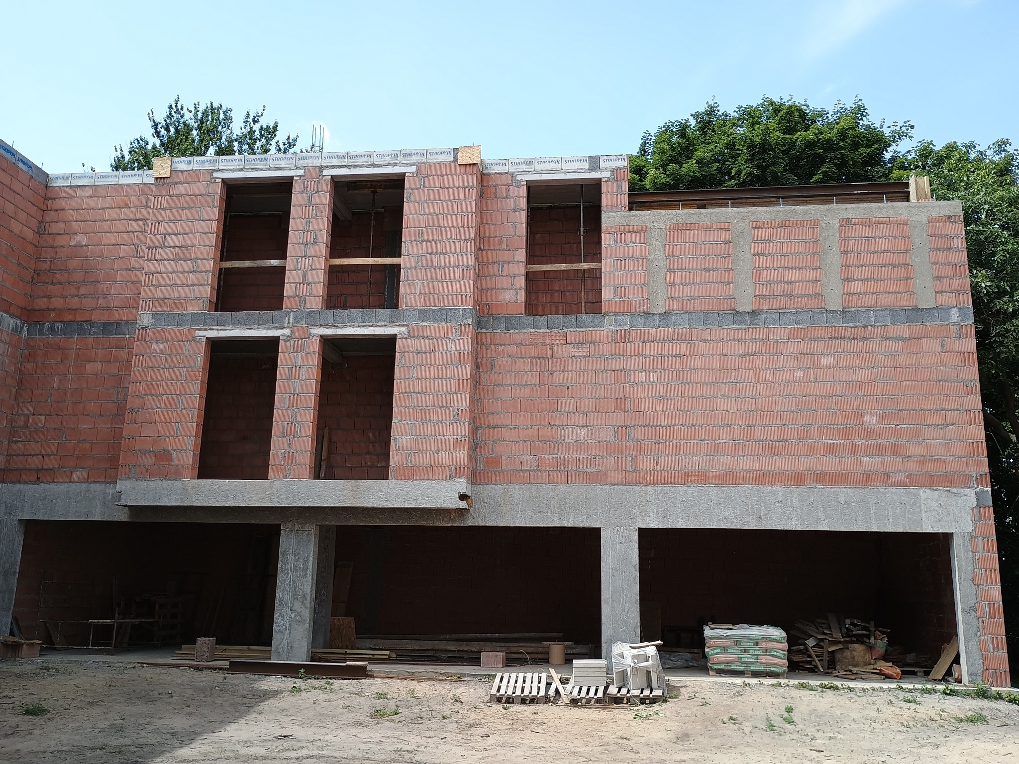 Dwukondygnacyjny budynek w trakcie budowy z czerwonej cegły, widoczne otwory okienne i betonowe nadproża, na dole otwarte przestrzenie garażowe, błękitne niebo w tle.
