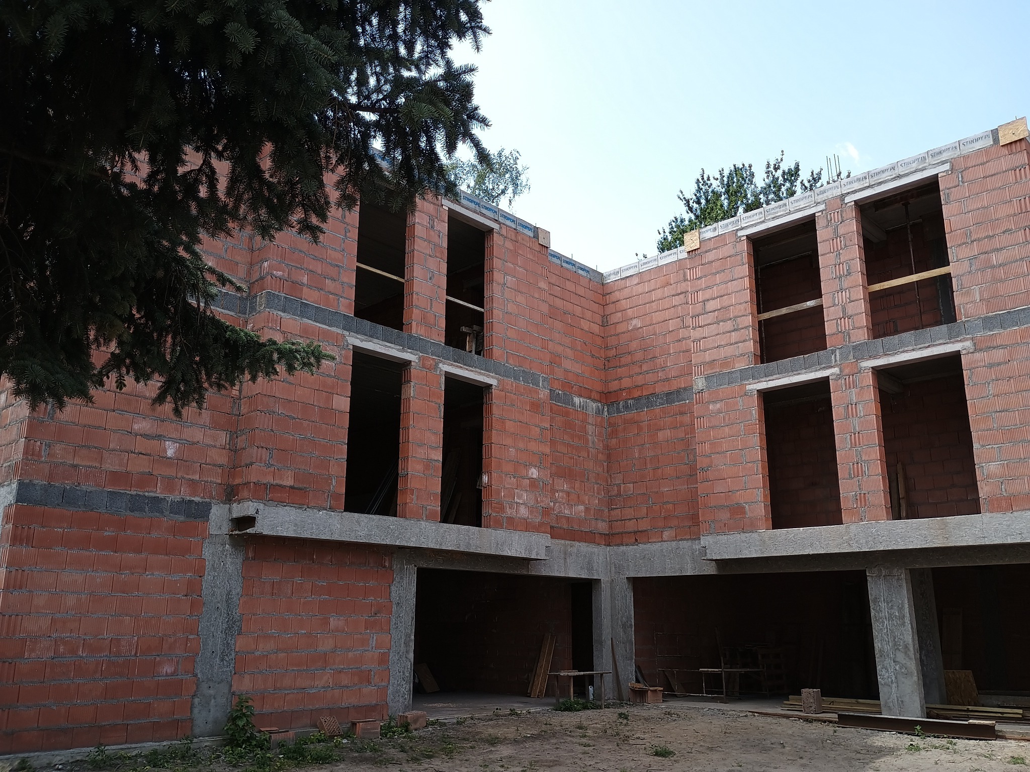Surowy stan domu z czerwonej cegły z widocznymi otworami okiennymi i betonowymi filarami, częściowo przysłonięty przez drzewo iglaste. Budowa w toku.